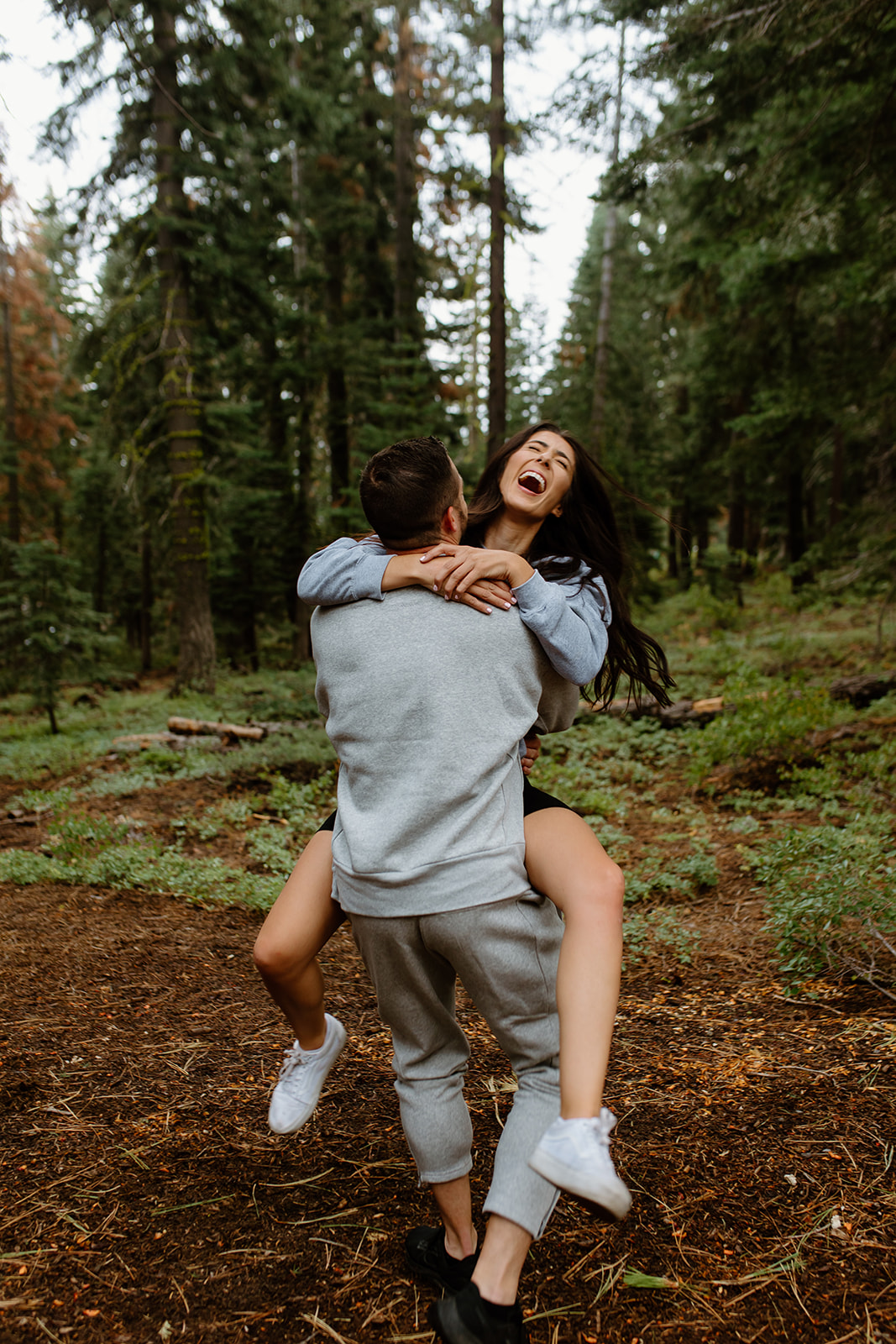 Brunette couple taking photos in the Lake Tahoe forest. They're both wearing grey cozy sweats and having fun laughing together while getting pictures taken by Wild Love & Wanderlust, a lake tahoe wedding photographer.