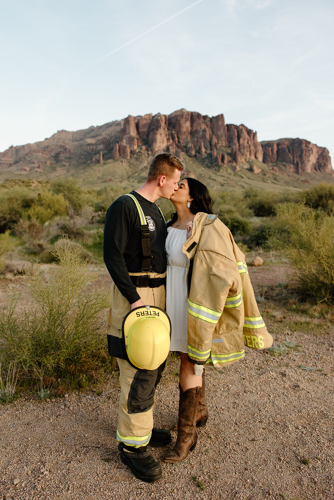 Lost Dutchman State Park, Blonde Man wearing firefighter uniform, his wife a brunette named Allie wearing a white dress and his firefighter jacket. The Superstition Mountains are behind them and it's sunset.